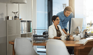 A person sitting at a desk and show another person something on a computer.