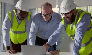 Three people looking at a laptop. Two of which are wearing hard hats.