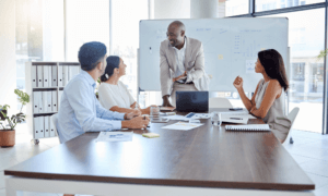 3 people sitting around a table looking at a colleague presenting a chart on the whiteboard.