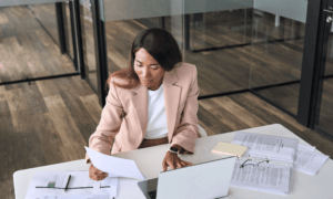 Someone working at a desk. They are looking at papers and typing on a laptop.