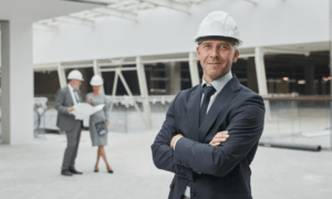 Construction worker smiling at the camera with two people discussing in the background.