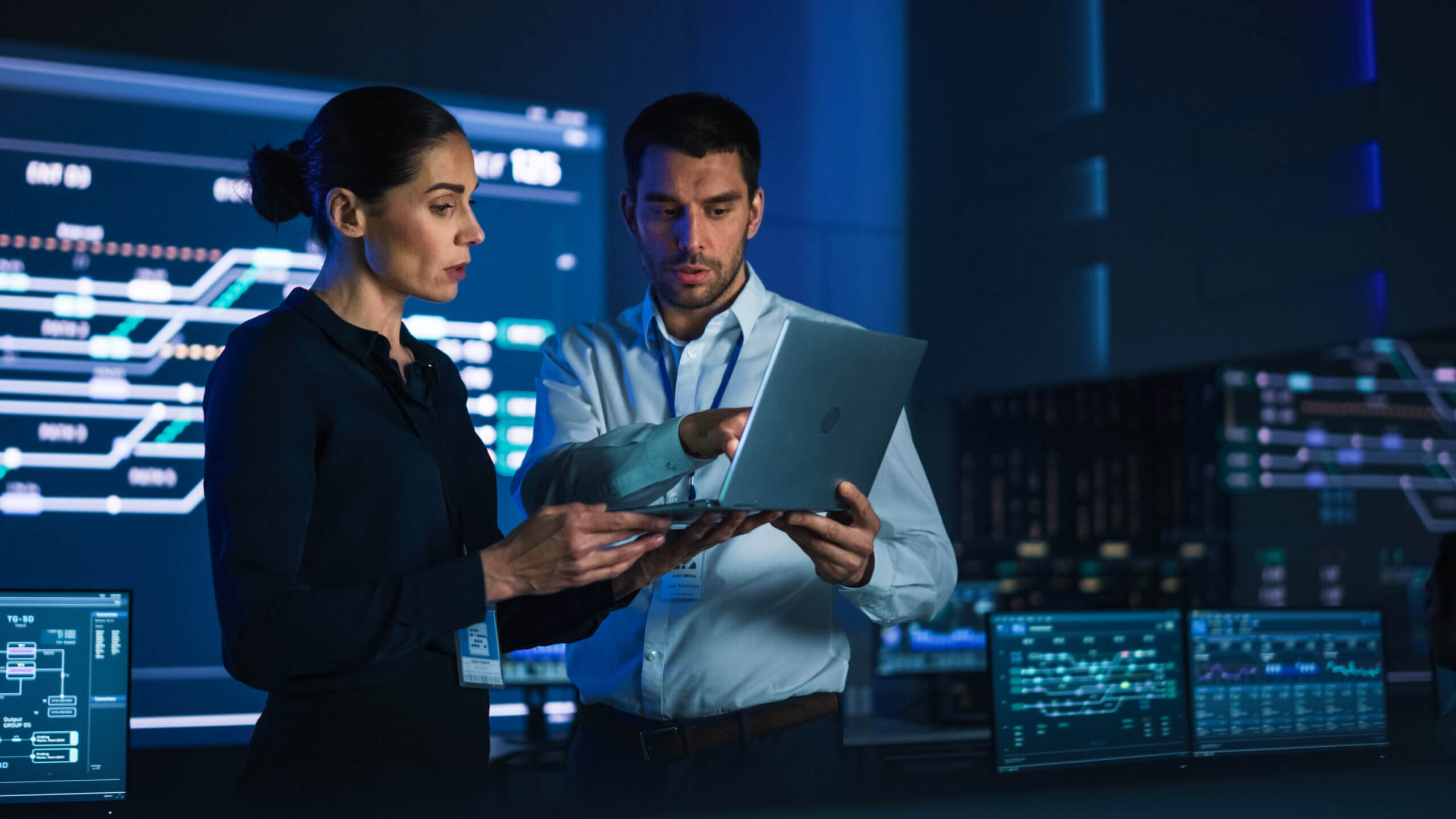 Female Project Manager works on Laptop Standing in Telecommunications System Control Room. In Background Big Screen Showing Infographics of Infrastructure, Charts, System Analysis