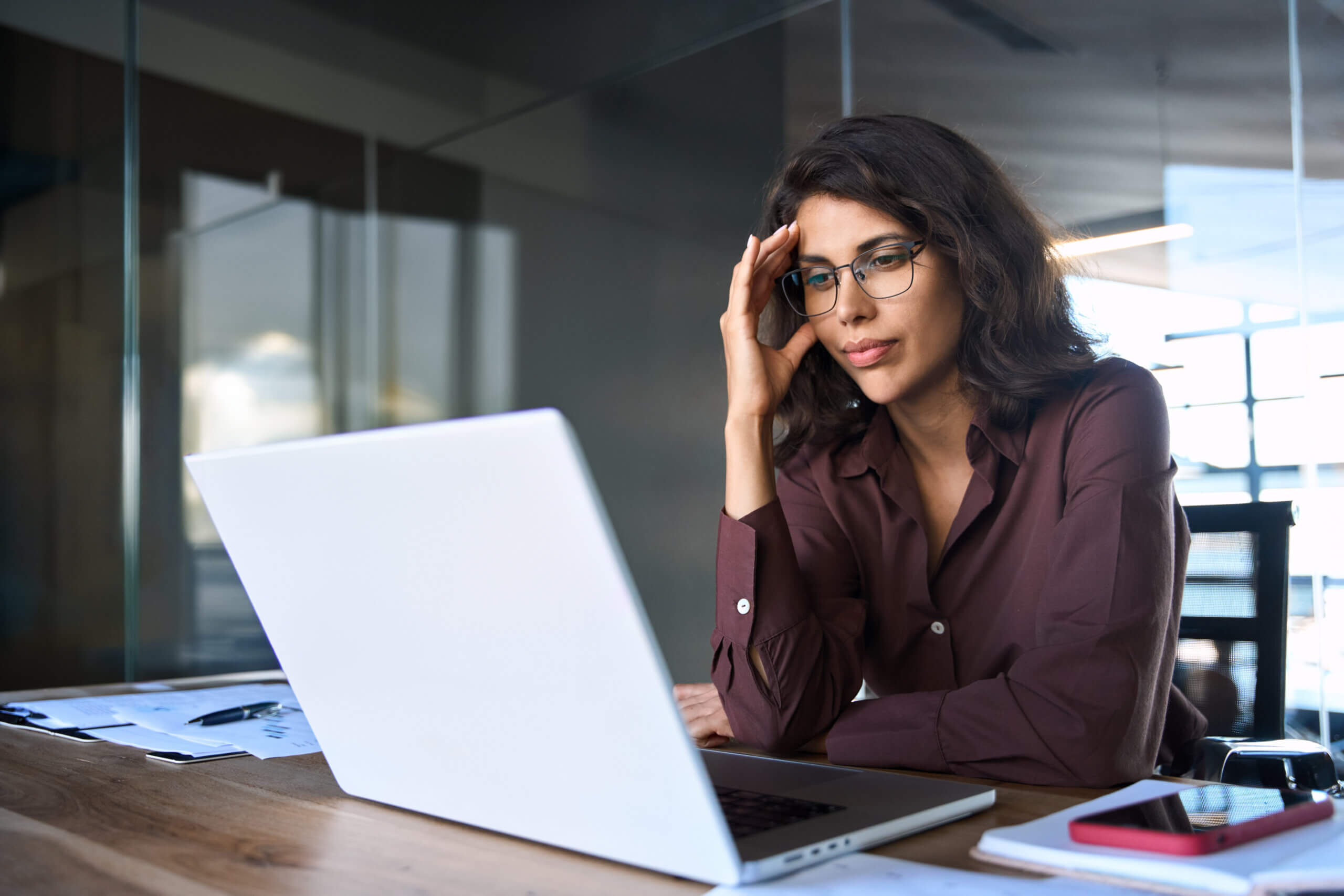 Focused young businesswoman looking at laptop pc computer screen having headache, migraine. Latin business woman holding hand near temples, feeling stressed and tired sitting at workplace in office